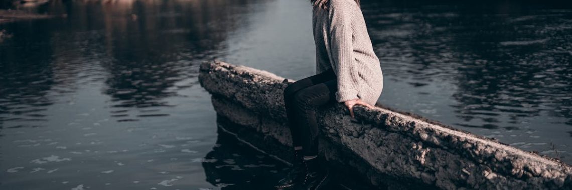 A woman enjoys a peaceful moment sitting on a riverside barrier during sunset.
